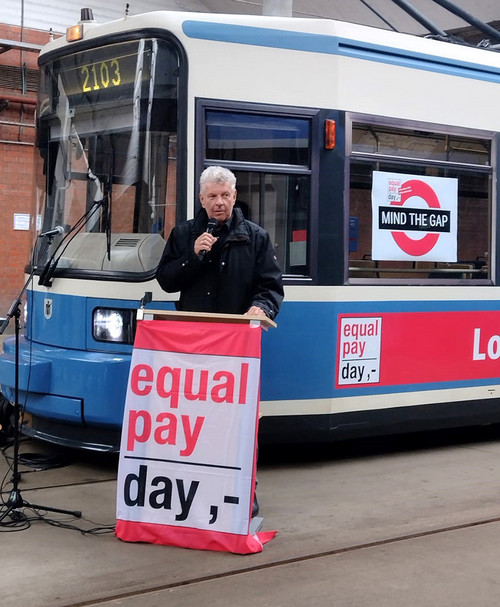Mann steht an einem Pult mit der Aufschrift "Equal Pay Day"