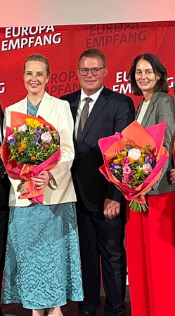 Zwei Frauen mit Blumensträußen, in der Mitte ein Mann.