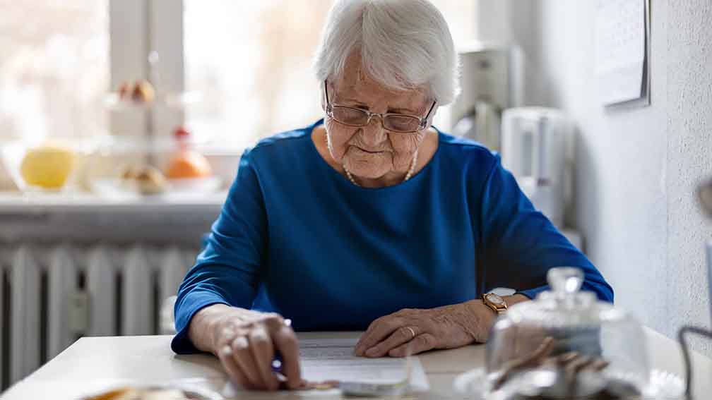 Ältere Frau sitzt am Tisch und hat einen Zettel vor sich. 