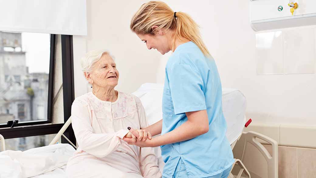 Pflegerin hält die Hand einer älteren Frau, die im Bett sitzt. 