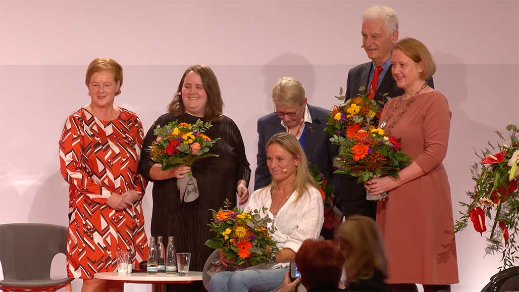 6 Personen mit Blumensträußen auf einer Bühne