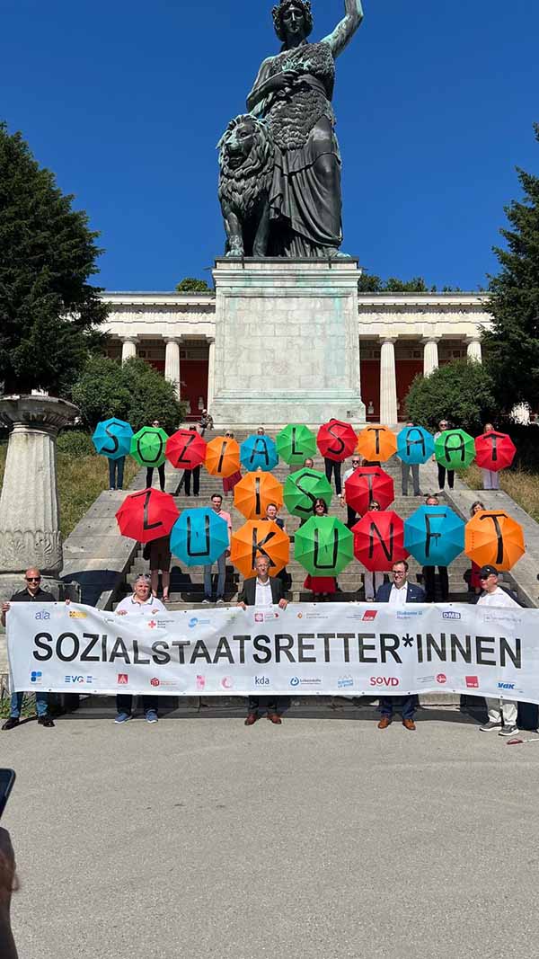 Viele Menschen stehen mit einem langen Plakat und Luftballons vor der Bavaria