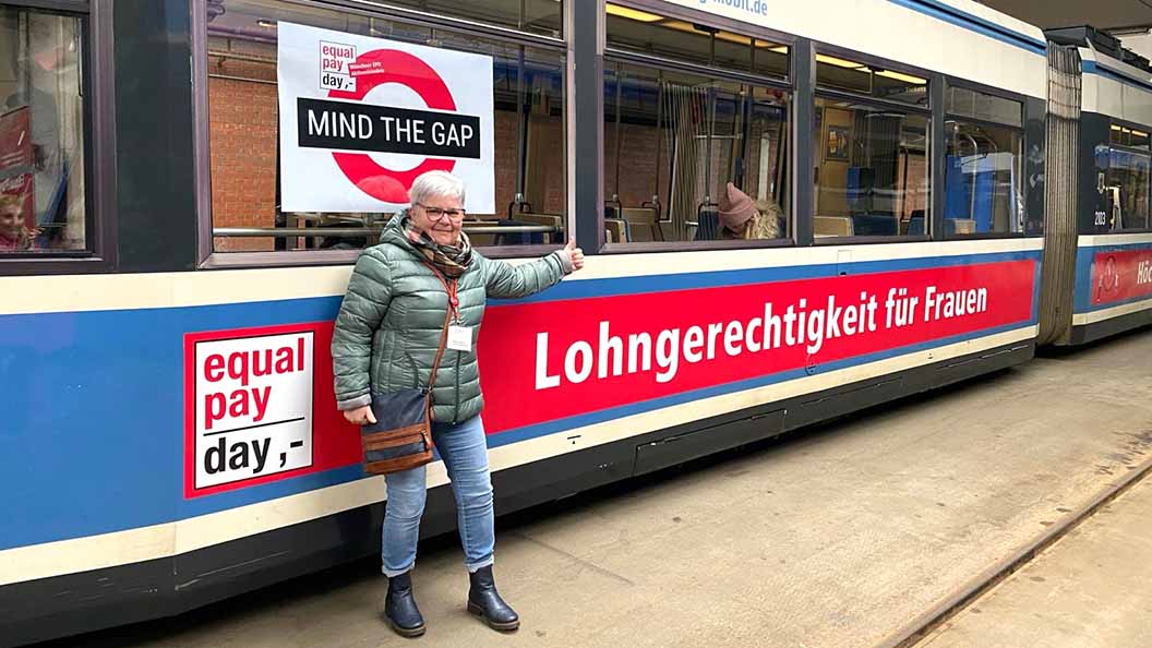 Frau steht vor einer Straßenbahn mit dem Aufdruck "Lohngerechtigkeit für Frauen"